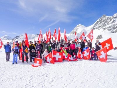 Group of people with Swiss flags in the snow, surrounded by snow-covered mountains.