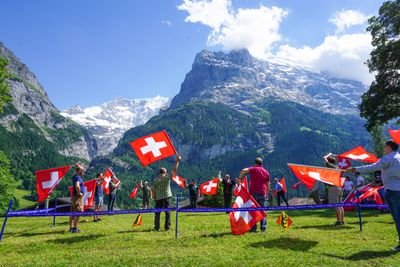 People are waving Swiss flags on a meadow in front of a mountain backdrop.