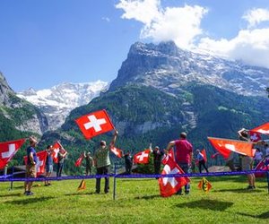 People are waving Swiss flags on a meadow in front of a mountain backdrop.
