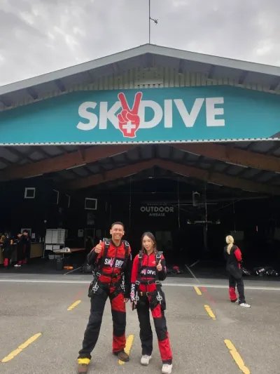 Two people in skydiving suits in front of a building with a "Skydive" sign.