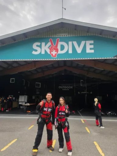 Zwei Personen in Fallschirmsprunganzügen vor einem Gebäude mit "Skydive"-Schild.