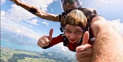 Two people tandem skydiving over a mountain landscape, wearing helmets and goggles.