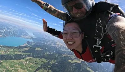 Two people tandem skydiving over a mountain landscape with a lake, wearing helmets and harnesses.