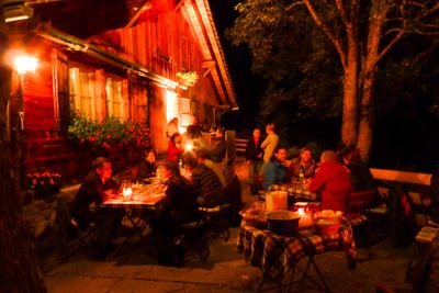 People are sitting at tables in front of a illuminated wooden house in the evening, surrounded by trees.