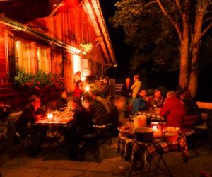 People are sitting at tables in front of a illuminated wooden house in the evening, surrounded by trees.