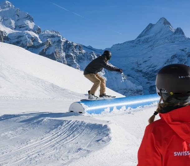 Snowboarder on rail in the snow park, observed by person in red jacket. Mountains in the background.