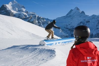 Snowboarder on rail in the snow park, observed by person in red jacket. Mountains in the background.