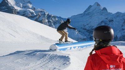 Snowboarder on rail in the snow park, observed by person in red jacket. Mountains in the background.