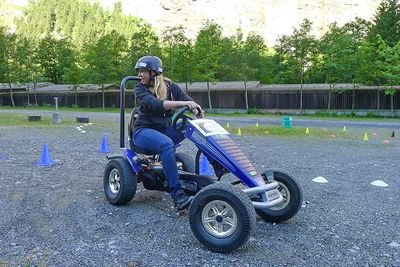 Person auf einem blauen Gokart mit Helm auf Schotterplatz, umgeben von Kegeln und Bäumen.