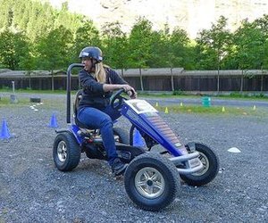 Person on a blue go-kart with a helmet on a gravel surface, surrounded by cones and trees.