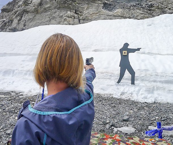 Person with jacket aiming a gun at a target in front of a snow-covered rock.