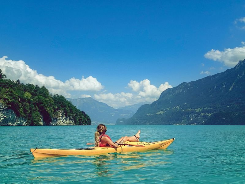 Person in a yellow kayak on a lake, surrounded by mountains and forest, under a clear sky.