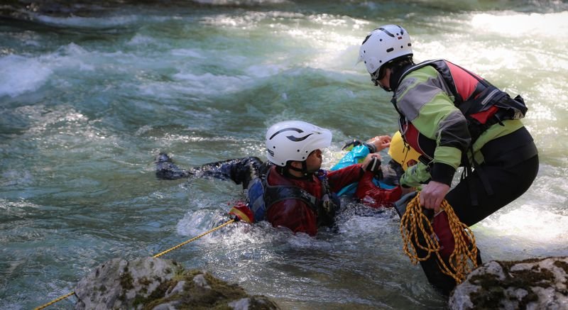 People with helmets and wetsuits canyoning in a river, one person with a rope helping.