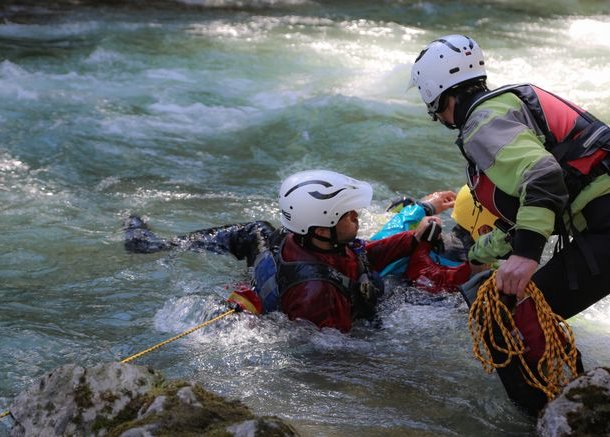 People with helmets and wetsuits canyoning in a river, one person with a rope helping.
