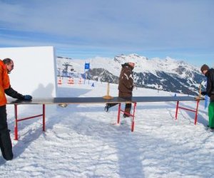 People are playing table curling on a snowy mountain wearing helmets and winter clothing.
