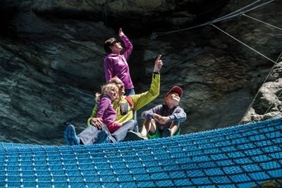 Personen auf blauem Netz in einer Felslandschaft, tragen Freizeitkleidung, zeigen nach oben.