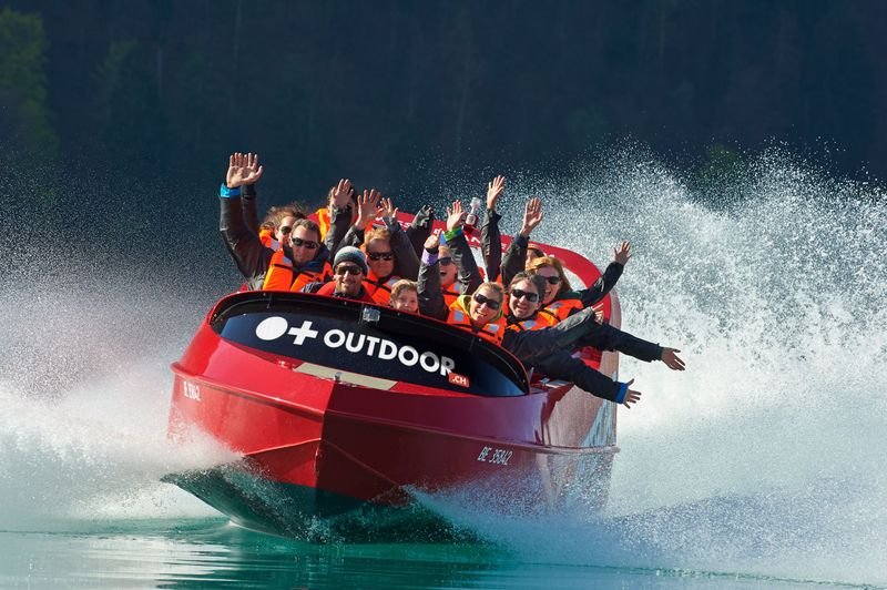 People in a red jet boat with life jackets on a lake, surrounded by splashing water.