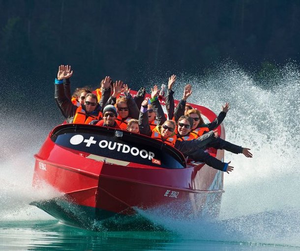People in a red jet boat with life jackets on a lake, surrounded by splashing water.