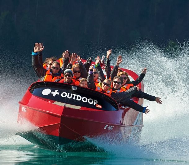 People in a red jet boat with life jackets on a lake, surrounded by splashing water.