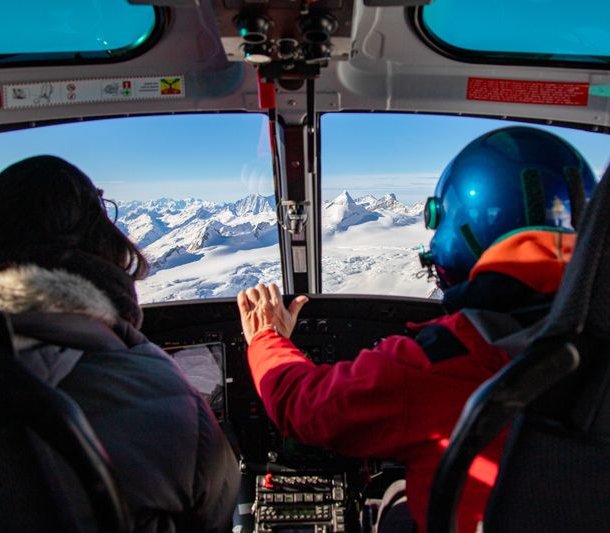 Two people in a helicopter cockpit, one wearing a blue helmet, flying over snowy mountains.