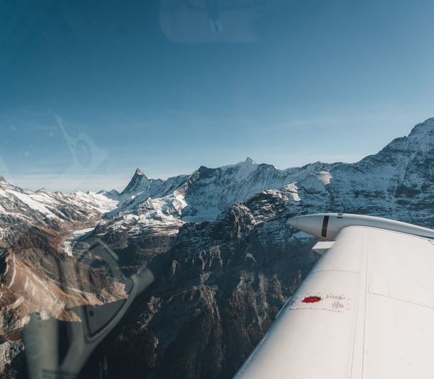 View from airplane wing over snow-capped mountains under clear blue sky.