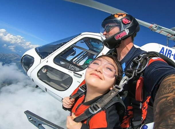 Zwei Personen, die aus einem Hubschrauber Fallschirmspringen, tragen Helme und Gurte, über Wolken und Bergen.