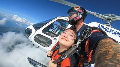 Two people skydiving from a helicopter, wearing helmets and harnesses, above clouds and mountains.