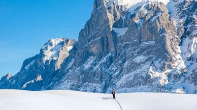 Person skiing with poles on snowy terrain, wearing a backpack, in front of large mountain peaks.