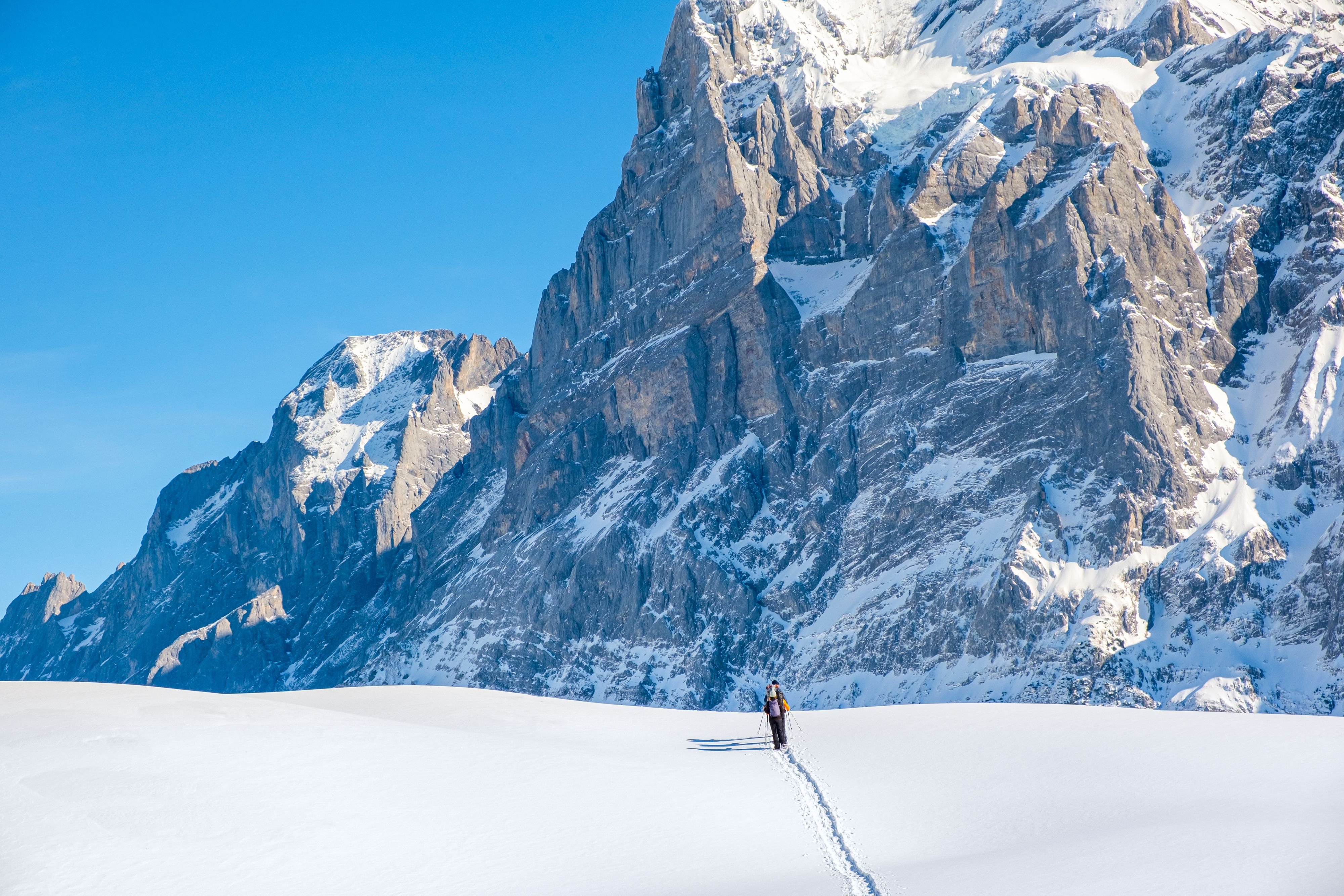 Person, der mit Stöcken auf schneebedecktem Gelände Ski fährt, mit einem Rucksack, vor großen Berggipfeln.