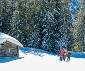 Drei Personen in Winterkleidung wandern mit Schneeschuhen in einem verschneiten Wald in der Nähe einer Holzhütte.