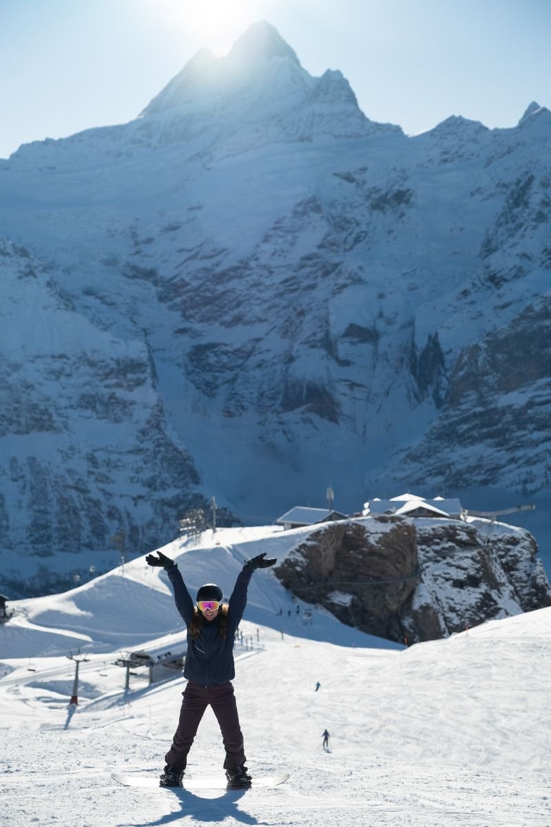 Person in winter gear with raised arms on snowy slope, mountains in background, clear sky above.