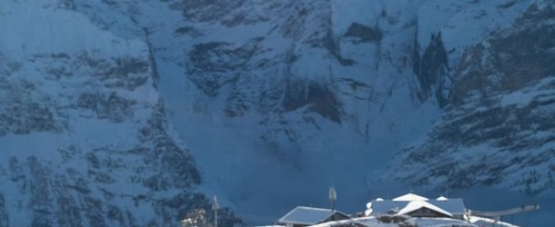 Person in winter gear with raised arms on snowy slope, mountains in background, clear sky above.