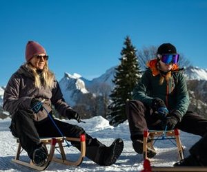 Two people sitting on sleds in snowy mountains, wearing winter clothing and gloves.