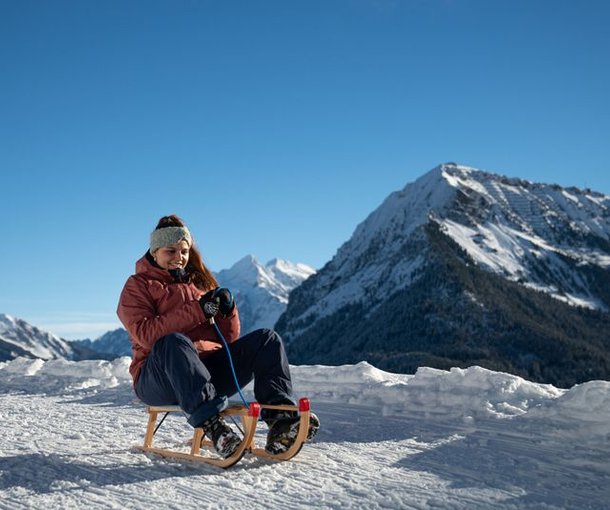 Person sledding on snow with a wooden sled, wearing a jacket, headband, and gloves; snowy mountains in the background.