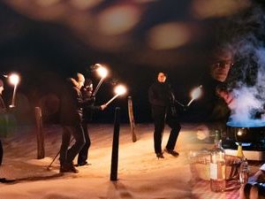 People with torches on a snowy path, next to a table with a steaming pot and bottles.