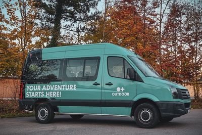 Ein türkisfarbener Van mit der Aufschrift "Your Adventure Starts Here!" steht vor herbstlichen Bäumen.