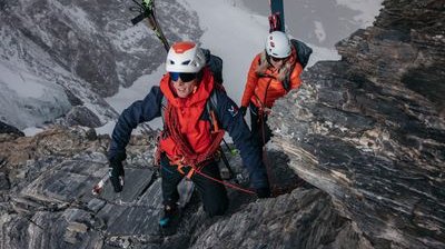 Zwei Personen klettern mit Helmen und Seilen auf felsigem Berg, Skier am Rucksack befestigt.