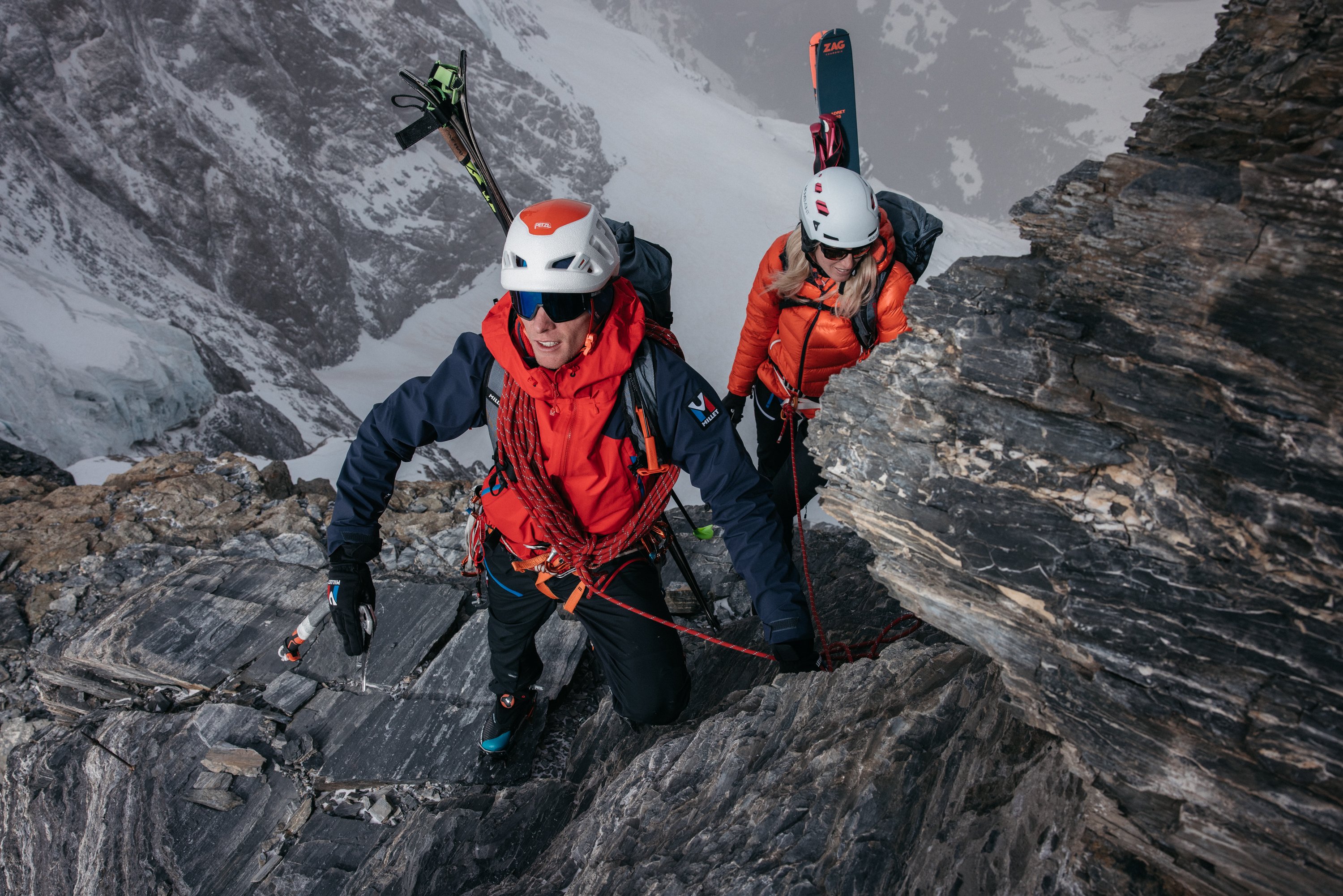 Zwei Personen klettern mit Helmen und Seilen auf felsigem Berg, Skier am Rucksack befestigt.