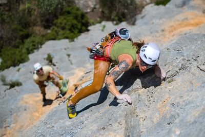 Zwei Kletterer mit Helmen und Gurten steigen an einer felsigen Klippe empor, umgeben von Grünzeug.