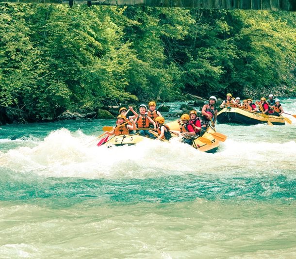 Groups rafting on a river, wearing helmets and life jackets, surrounded by lush green trees.