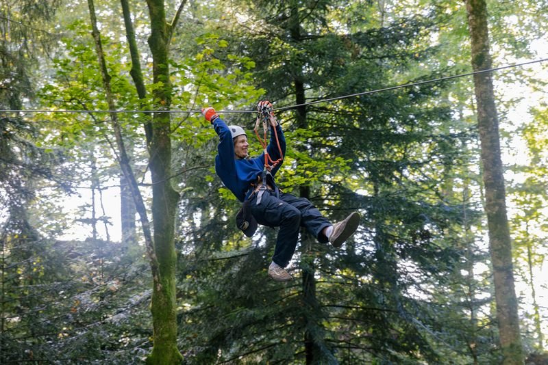 Person riding a zipline through the forest is wearing a helmet, a safety harness, and gloves, surrounded by trees.