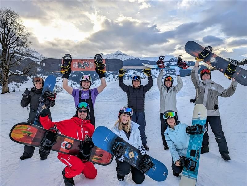 Group of people in winter gear holding snowboards overhead on a snowy mountain slope.