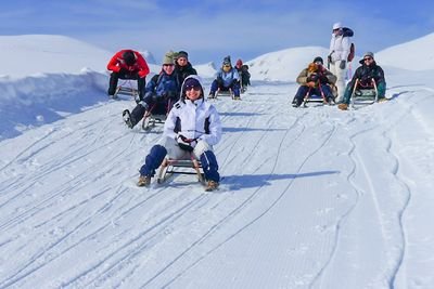 Personen rodeln auf einer verschneiten Piste in den Bergen, tragen Winterkleidung und Helme.
