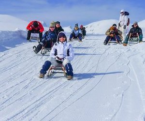 People are sledding on a snowy slope in the mountains, wearing winter clothes and helmets.