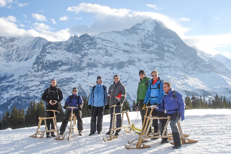 Eine Gruppe von Personen mit Schlitten auf schneebedecktem Berg vor alpiner Kulisse.