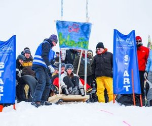 People in winter clothing and helmets start a sled race in the snow between blue banners.