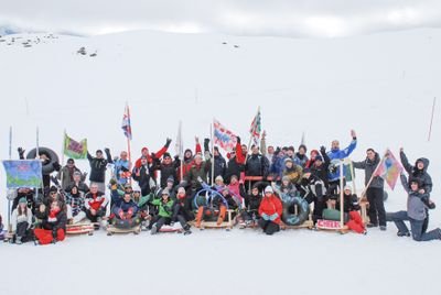 Group of people in the snow with sleds and flags, surrounded by snow-covered mountains.
