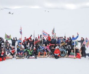 Group of people in the snow with sleds and flags, surrounded by snow-covered mountains.
