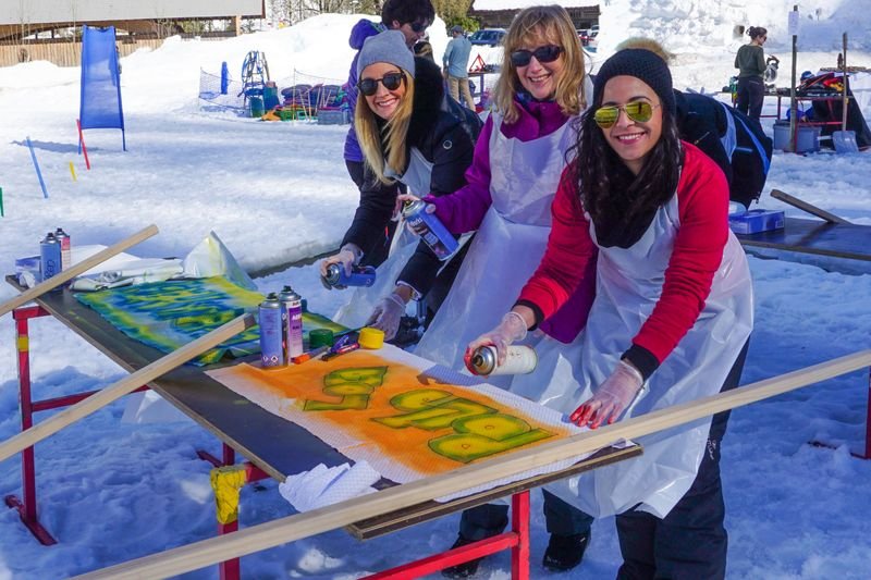 Drei Personen sprühen Graffiti auf Holzplatten im Schnee, tragen Schürzen und Handschuhe.