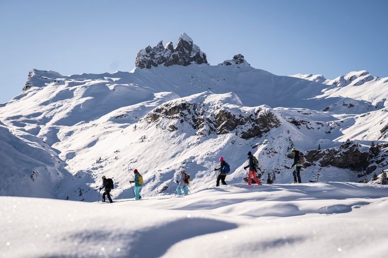 People with skiing equipment are hiking through a snowy mountain landscape under a clear sky.